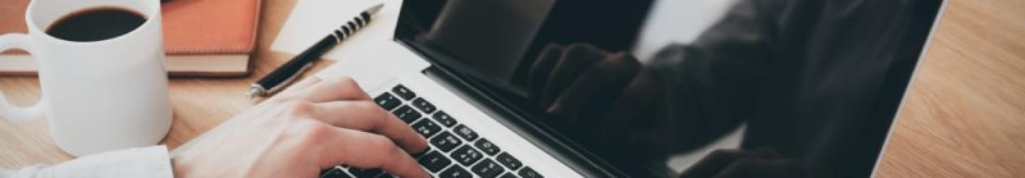 zwei Hände auf einer Laptop-Tastatur, daneben eine Kaffeetasse und Arbeitsutensilien